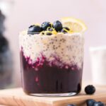 A glass of blueberry lemon overnight oats topped with blueberries, lemon zest, and lemon slice. Blueberries scattered around.