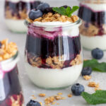 A jar of blueberry parfait topped with fresh mint and blueberries with more jars around it and toppings scattered around.