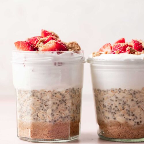 Two jars of cheesecake overnight oats topped with chopped strawberries and crumbled graham crackers.