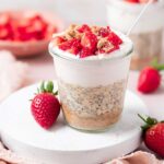 A jar of cheesecake overnight oats topped with chopped strawberries and crushed graham crackers. Fresh strawberries scattered around.