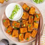 Overhead view of a plate of air fryer tofu garnished with green onions with a bowl of sauce on the side with more green onions.