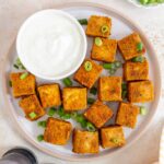 Overhead view of a plate of super crispy oven baked tofu garnished with green onions with a bowl of sauce on the side.