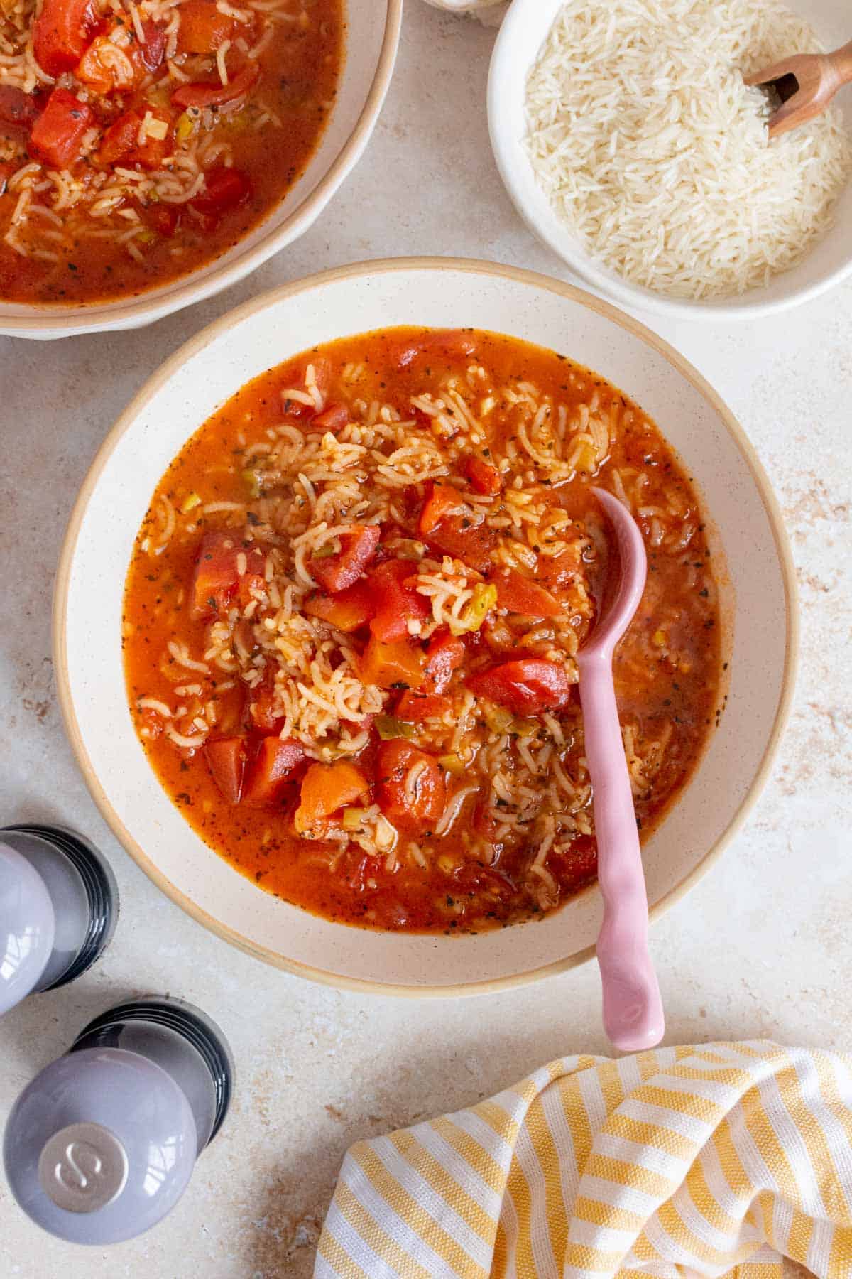 Overhead view of a bowl of tomato rice soup with a spoon. A towel, salt and pepper shakers, a second bowl of soup, and some rice off to the side.