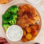 A close up view of a plate with an oven baked bone-in pork chop with potatoes, broccoli, and a bowl of herby sauce.