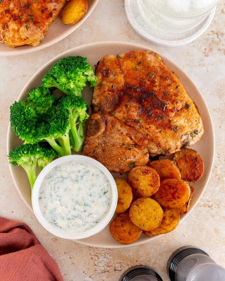 A close up view of a plate with an oven baked bone-in pork chop with potatoes, broccoli, and a bowl of herby sauce.