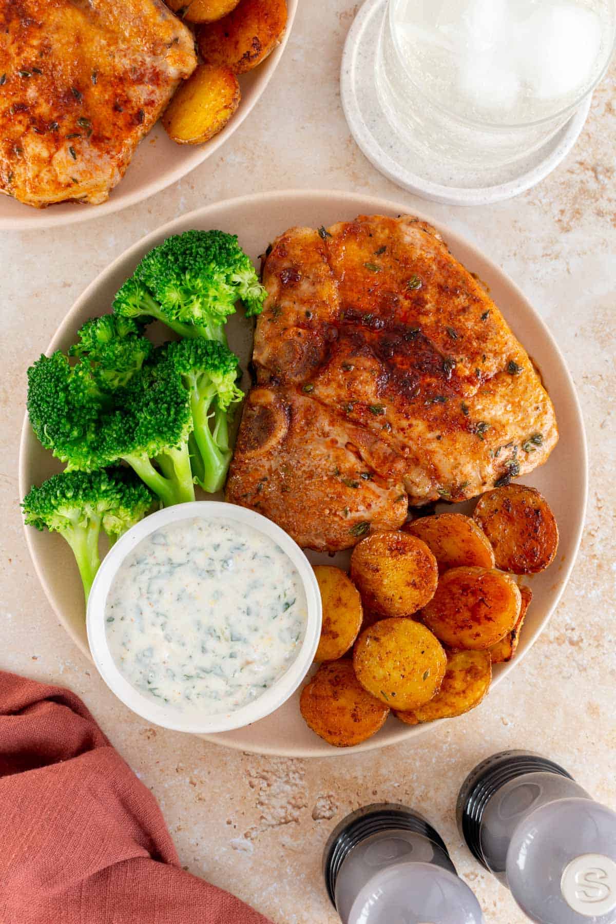A close up view of a plate with an oven baked bone-in pork chop with potatoes, broccoli, and a bowl of herby sauce.