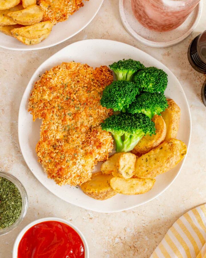 An overhead view of a plate with a baked chicken cutlet along with broccoli and potato wedges. Another plate, a drink, and dip off to the side.