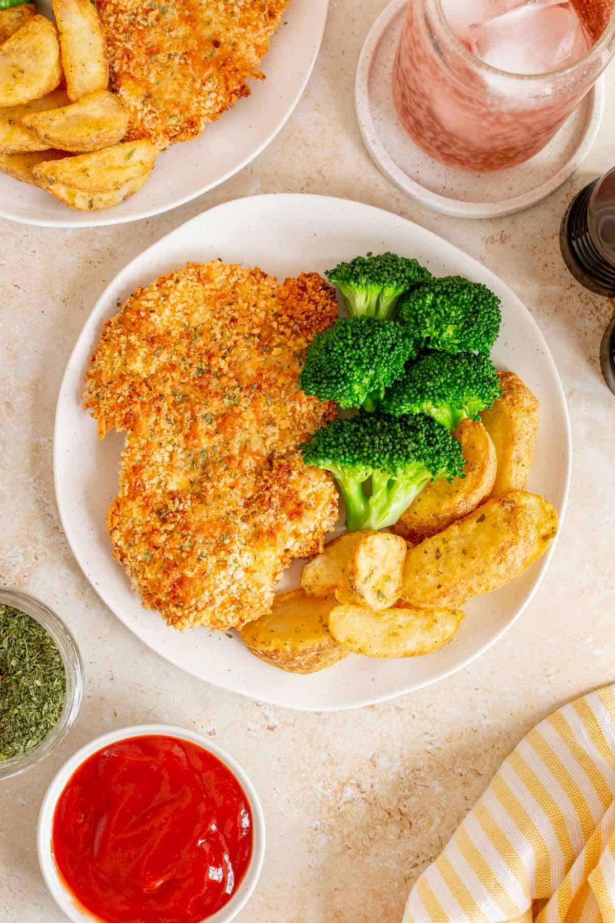 An overhead view of a plate with a baked chicken cutlet along with broccoli and potato wedges. Another plate, a drink, and dip off to the side.