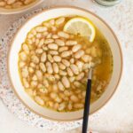 An overhead view of a bowl of brothy beans (white beans recipe) with a lemon slice.