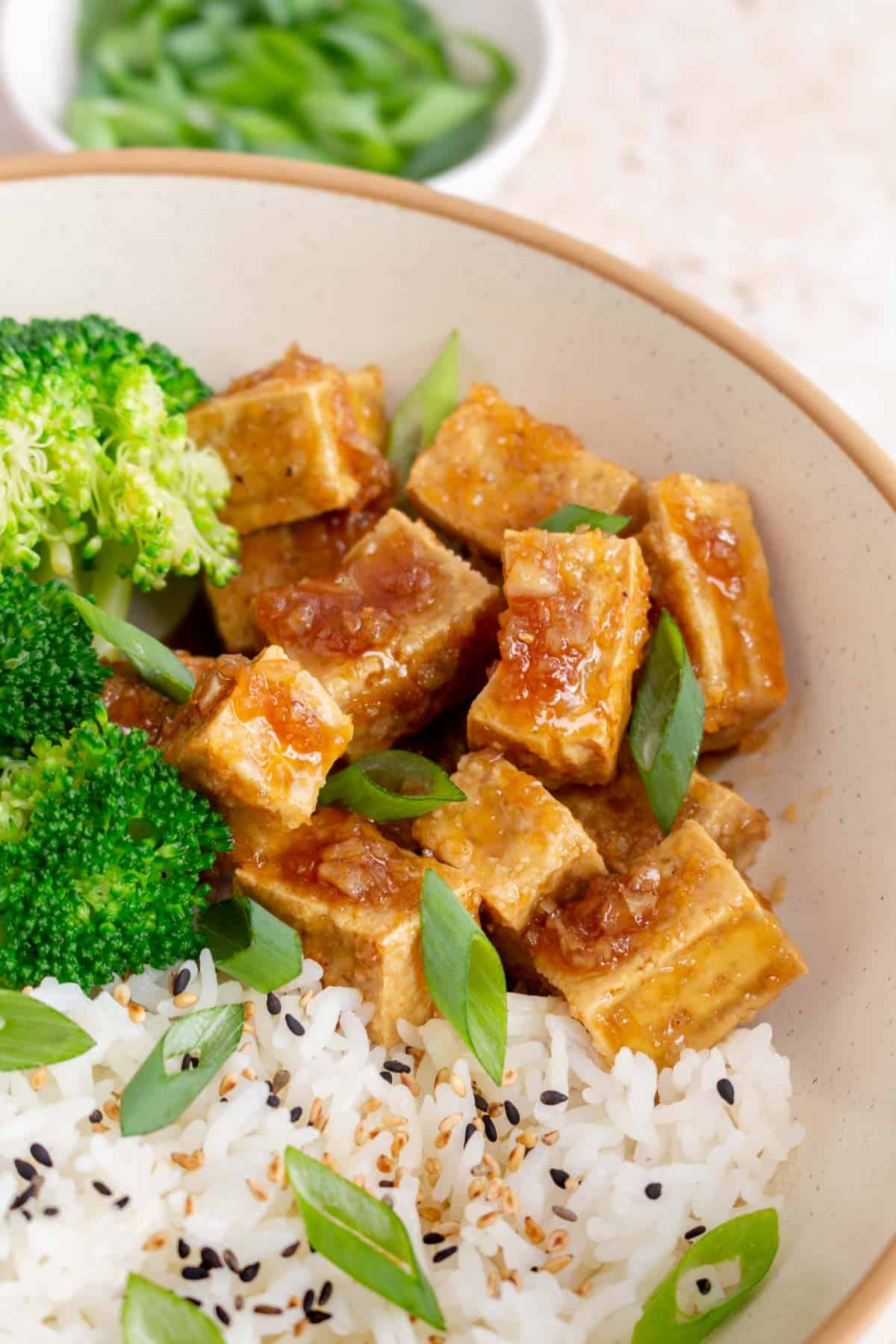 A close up view of honey garlic tofu in a bowl with rice, broccoli, and green onions.