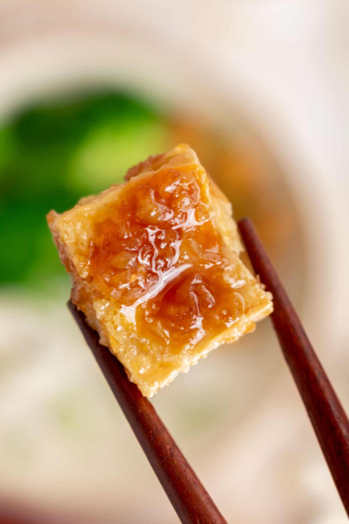 A close up view of honey garlic tofu held up by a pair of chopstick.