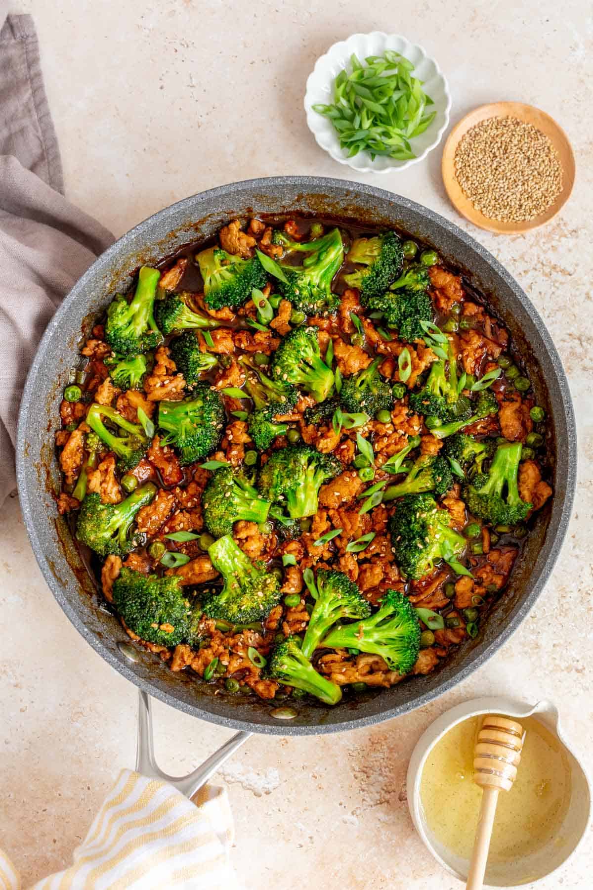 An overhead view of a skillet of honey sriracha ground chicken and broccoli. Garnishes and honey on the side.