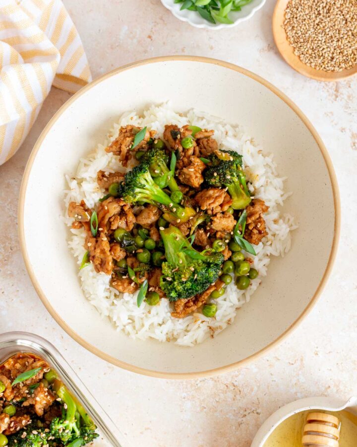 A bowl of honey sriracha ground chicken and broccoli over rice. Garnishes, a meal prep container, and bowl of honey on the side.
