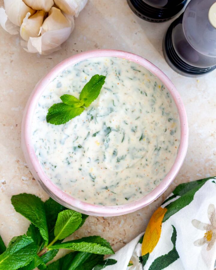 Overhead view of a bowl of mint yogurt sauce with fresh mint leaves as garnish. Fresh mint, garlic, linen, and salt and pepper shakers on the side.