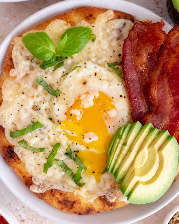 A plate with naan breakfast pizza with the egg yolk running. Basil, bacon, and avocado on top.