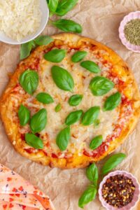 Overhead view of a naan pizza topped with fresh basil and surrounded by red pepper flakes, parmesan, and dried oregano in bowls.