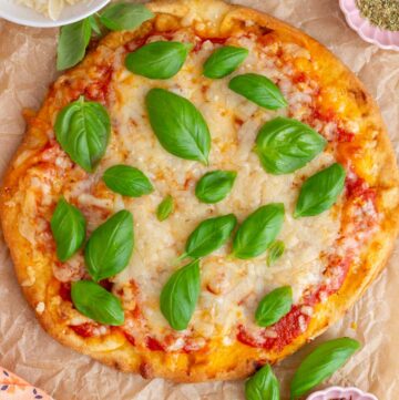 Overhead view of a naan pizza topped with fresh basil and surrounded by red pepper flakes, parmesan, and dried oregano in bowls.