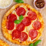 An overhead view of a pepperoni flatbread pizza with a basil garnish. A bowl of shredded cheese, red pepper flakes, and dried oregano on the side.