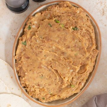 A platter of refried beans surrounded by half a lime, a linen napkin, tortillas, and salt and pepper shakers.
