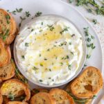 A bowl of whipped ricotta topped with pepper, thyme, honey, and lemon zest on a platter with toasted sliced baguette.
