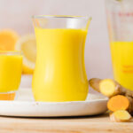 A ginger turmeric shot on a plate surrounded by cut ginger and turmeric and more juice.