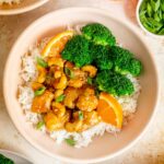 Overhead view of a bowl of rice with orange cashew chicken on top along with broccoli florets and orange slices.