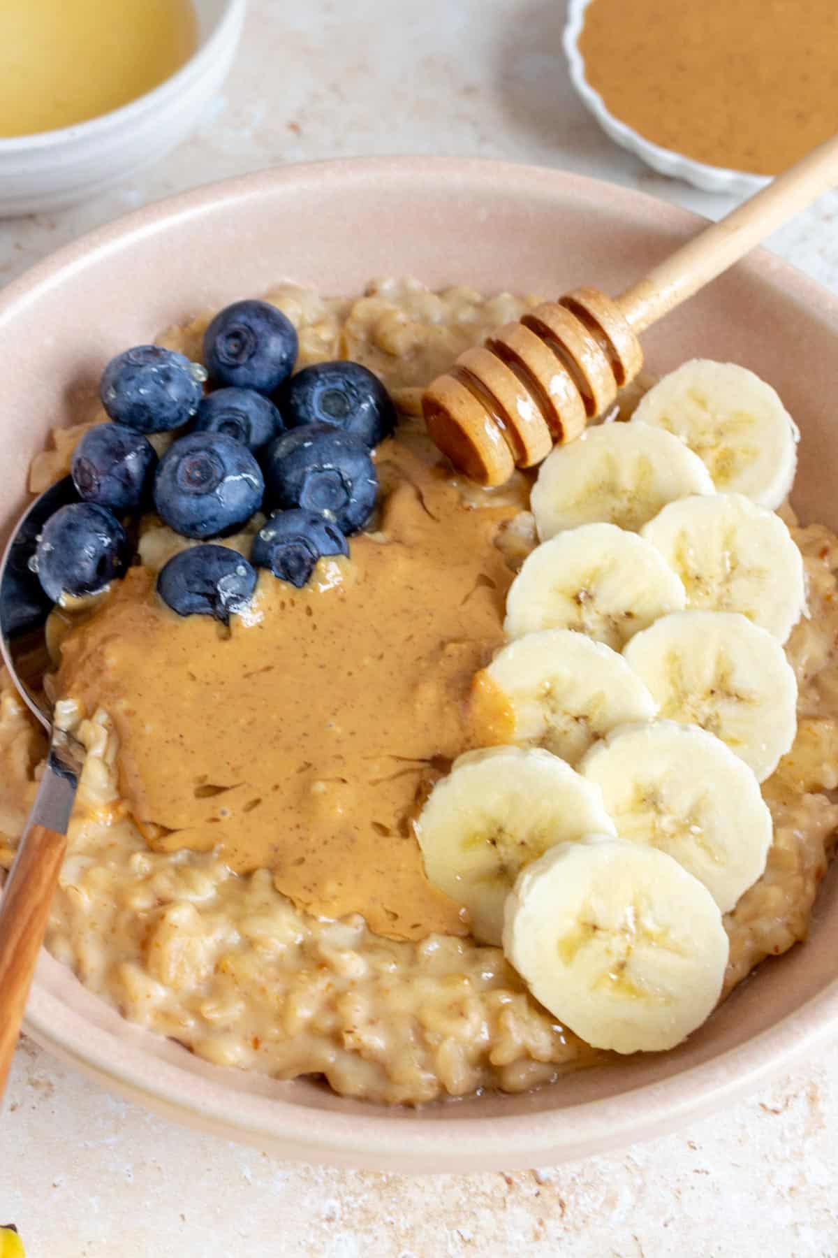 A bowl of peanut butter oatmeal topped with sliced bananas, peanut butter, and blueberries with a spoon tucked in and wooden honey dipper placed on top.