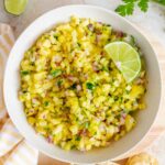 Overhead view of a bowl of pineapple salsa with a lime wedge on top. Tortilla chips, limes, and cilantro on the side.