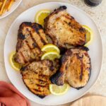 Overhead view of a platter with four grilled Italian pork chops with lemon slices.