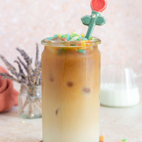 A glass of iced cereal milk latte with fruity pebbles on top.