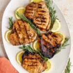 A platter with four grilled rosemary pork chops with lemon slices and rosemary.
