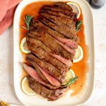 Overhead view of a grilled flank steak sliced in a platter with lemon slices and rosemary.