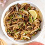 Overhead view of a plate of sautéed mushrooms and onions with green onion garnish and a slice of lemon.