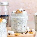 A jar of s'mores overnight oats on a wooden board surrounded by toppings and more jars off to the side.