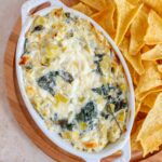 A small oval baking dish of Greek yogurt spinach artichoke dip on a wooden serving board with tortilla chips.