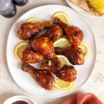 A platter of air fryer bbq chicken drumsticks with lemon slices between. A bowl of bbq, a plate of corn, and salt and pepper shakers on the side.