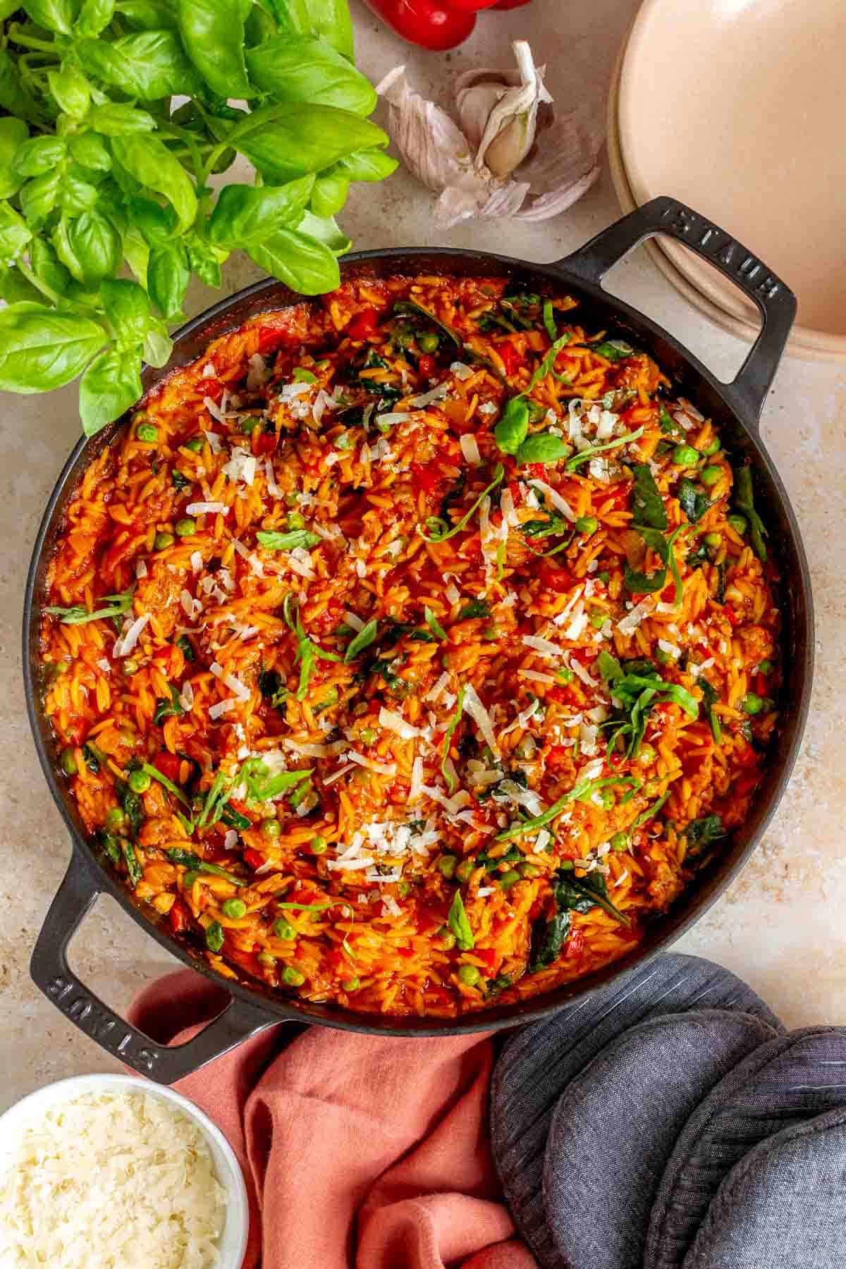 Overhead view of a large skillet of sausage orzo topped with shredded parmesan and basil.