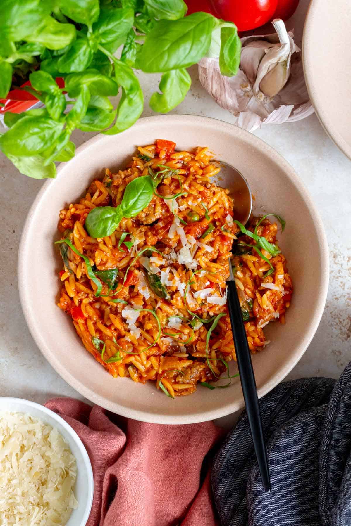 A pink bowl of sausage orzo topped with basil with a spoon inside.