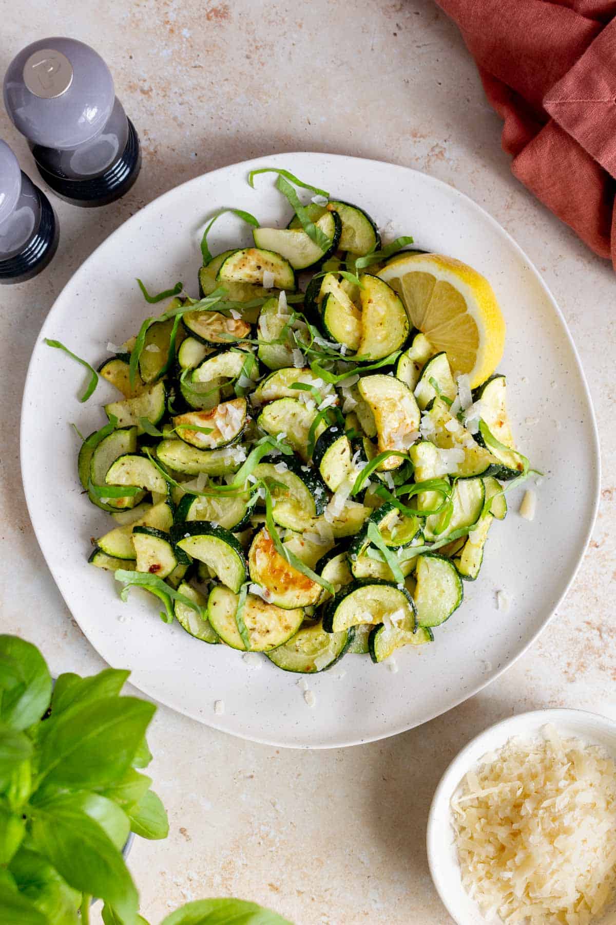 Overhead view of a platter of roasted zucchini with a wedge of lemon, garnished with basil ribbons and grated parmesan.