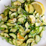 A close up view of a plate of roasted zucchini topped with basil and parmesan with a wedge of lemon.