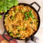 An overhead view of a large skillet of chicken broccoli orzo garnished with basil. Basil, a bowl of shredded cheese, salt, and pepper on the side.