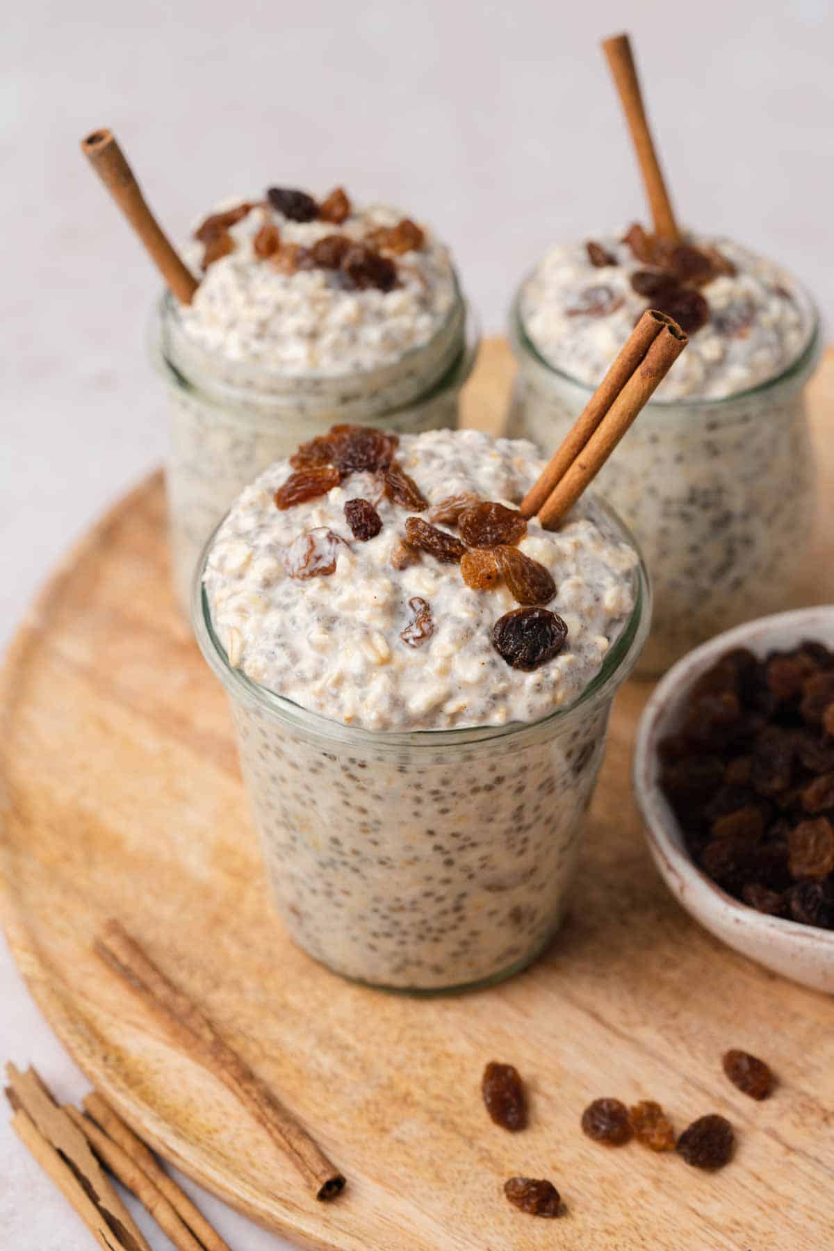 Three jars of cinnamon raisin overnight oats all topped with raisins and with a cinnamon stick stuck inside.