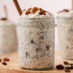 A profile view of a jar of cinnamon raisin overnight oats topped with raisins and cinnamon stick stuck inside.