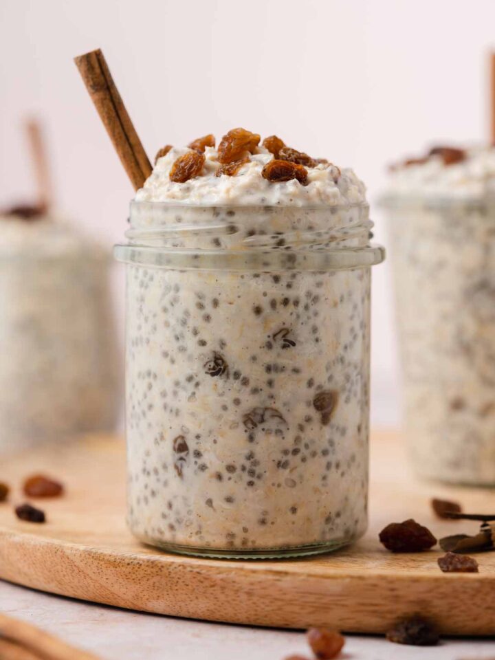 A profile view of a jar of cinnamon raisin overnight oats topped with raisins and cinnamon stick stuck inside.