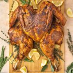 Overhead view of a wooden serving board with a dry brined spatchcock turkey, uncut, surrounded by fresh sage, thyme, rosemary, and lemon slices.