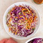 Overhead view of a large plate of asian coleslaw. A bowl of salad dressing, toasted sesame seeds, napkin, and another bowl of salad on the side.