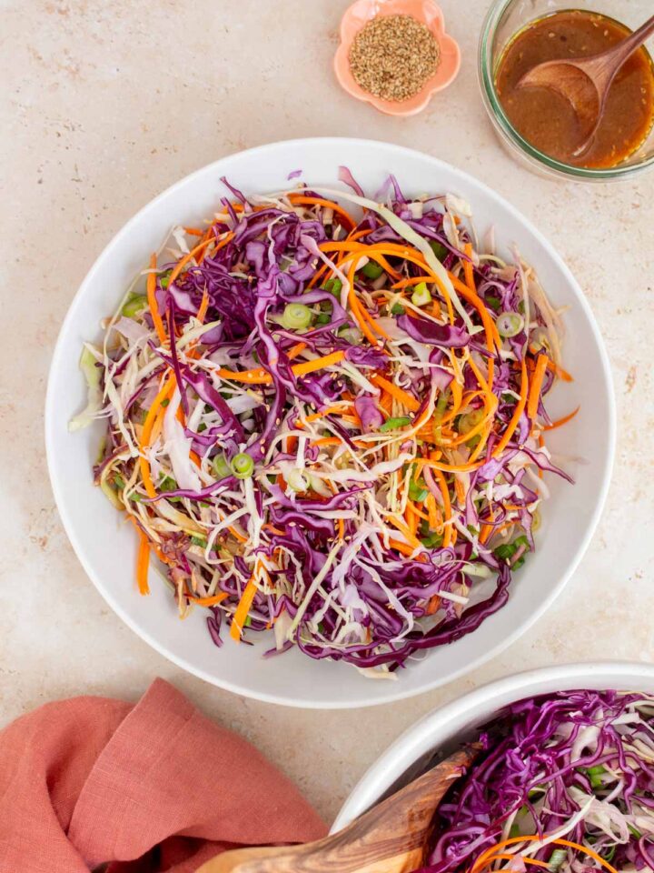 Overhead view of a large plate of asian coleslaw. A bowl of salad dressing, toasted sesame seeds, napkin, and another bowl of salad on the side.
