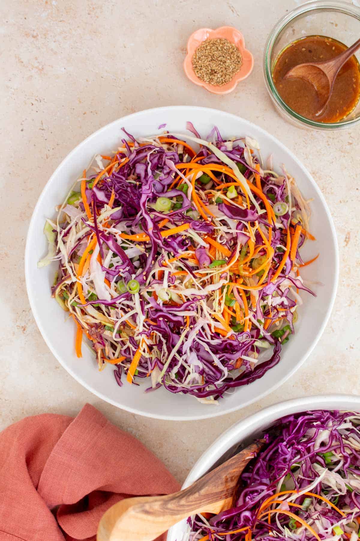 Overhead view of a large plate of asian coleslaw. A bowl of salad dressing, toasted sesame seeds, napkin, and another bowl of salad on the side.