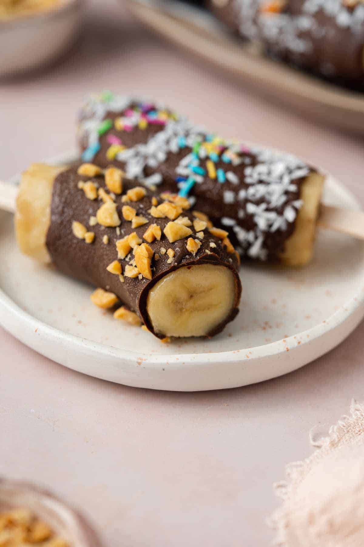 A close up view of two chocolate covered banana popsicles, one topped with roasted chopped peanuts and the other with sprinkles with a bite taken out of them.