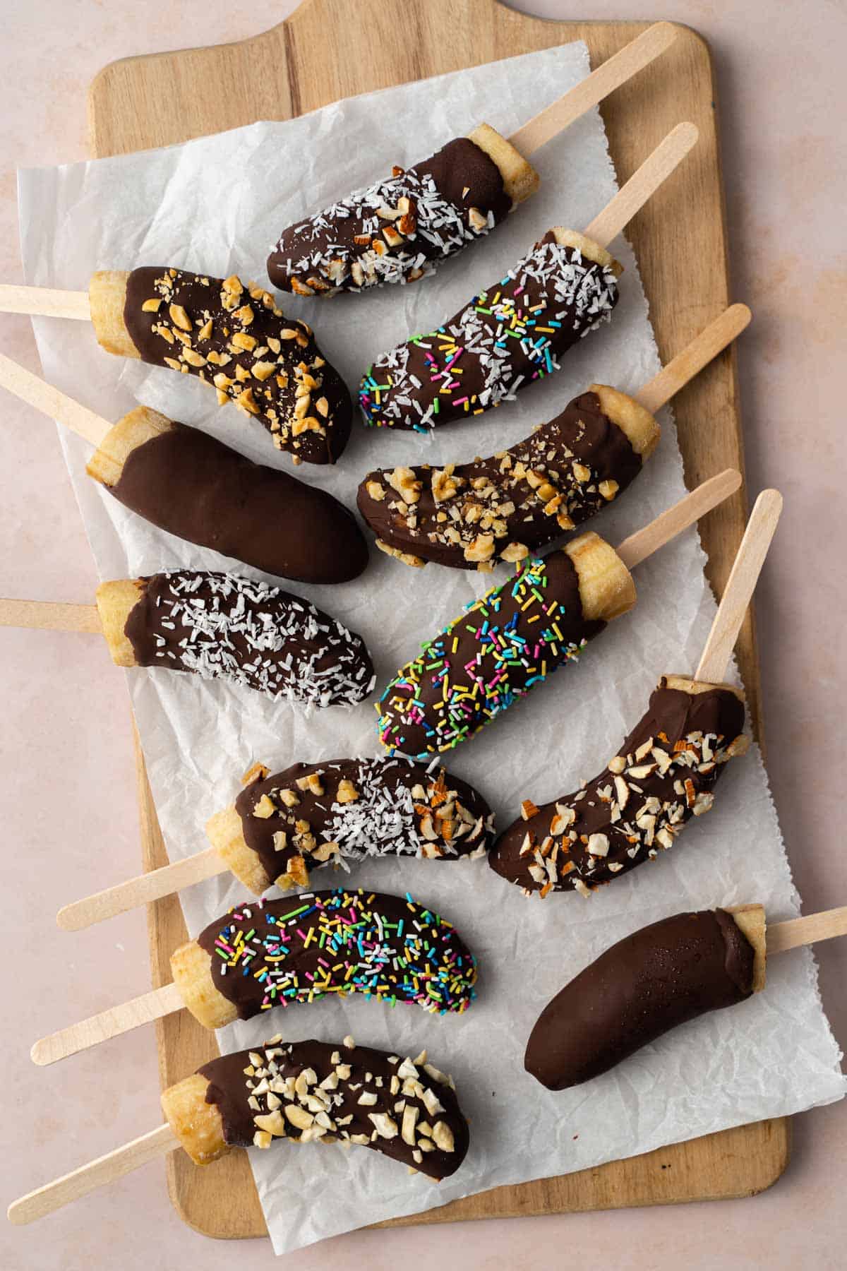A parchment lined wooden serving board with chocolate covered banana popsicles with various toppings.